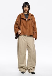 Bruine suedew jacket met franjes, gedragen over een zwart graphic t-shirt, gecombineerd met brede beige broeken en zwarte schoenen.