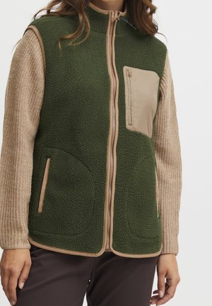 Kvinde iført grøn fleecevest med beige kant og brystlomme over beige striktrøje og mørke bukser.