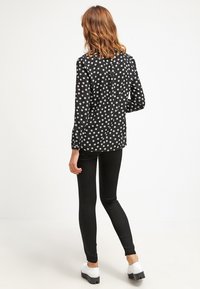 Blusa nera con motivo floreale bianco, maniche lunghe, bottoni sulla schiena, indossata con leggings neri e scarpe platform bianche.