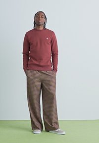 Bordeauxkleurige sweatshirt met geribbelde manchetten, gecombineerd met een brede, geruite broek in bruin en roomkleur. De schoenen zijn lichtgekleurde sneakers.