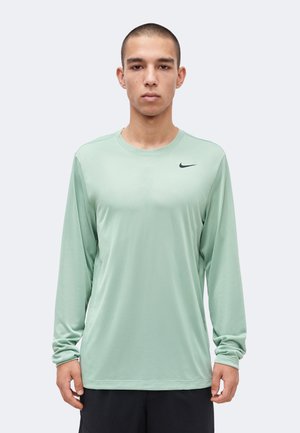 Tânăr cu păr scurt purtând un tricou verde deschis cu mâneci lungi Nike și pantaloni negri, privind înainte pe un fundal simplu.