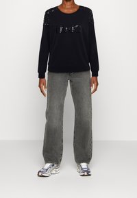 Sudadera negra con acentos de lentejuelas en los hombros, con el logo "LIU JO". Combinada con jeans de pierna ancha grises y zapatillas multicolores.
