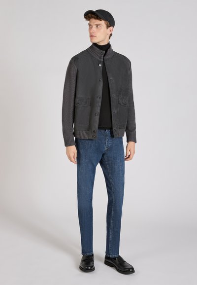 Grijze jasje met knopen en een gebreide kraag, gecombineerd met donkere spijkerbroek en zwarte schoenen. Model draagt een zwarte pet en toont gelaagd textuur.