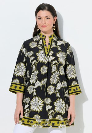 Frau trägt eine schwarze Tunika mit weißem Blumenmuster und olivgrünem Besatz mit kreisförmigen Mustern, stehend vor einem weißen Hintergrund.