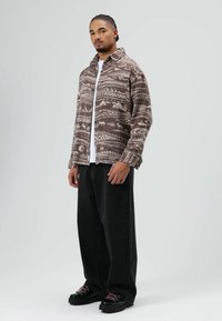 Bruine blousejas met patroon, een kraag, knopen aan de voorkant en lange mouwen, gecombineerd met zwarte jeans met wijde pijpen en dikke zwarte schoenen.