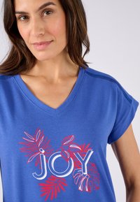 Camiseta de algodón azul con cuello en V que presenta el texto "JOY" en rojo y blanco, acompañado de ilustraciones decorativas de hojas. Mangas cortas y corte relajado.