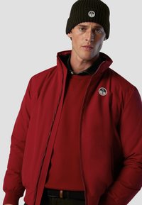 Giacca rossa con zip e colletto alto, texture morbida e toppa con logo; abbinata a un maglione rosso coordinato e a un berretto scuro.