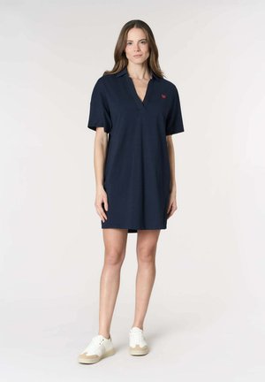 Donna in piedi che indossa un abito polo a maniche corte blu navy e scarpe da ginnastica bianche su uno sfondo chiaro e uniforme.