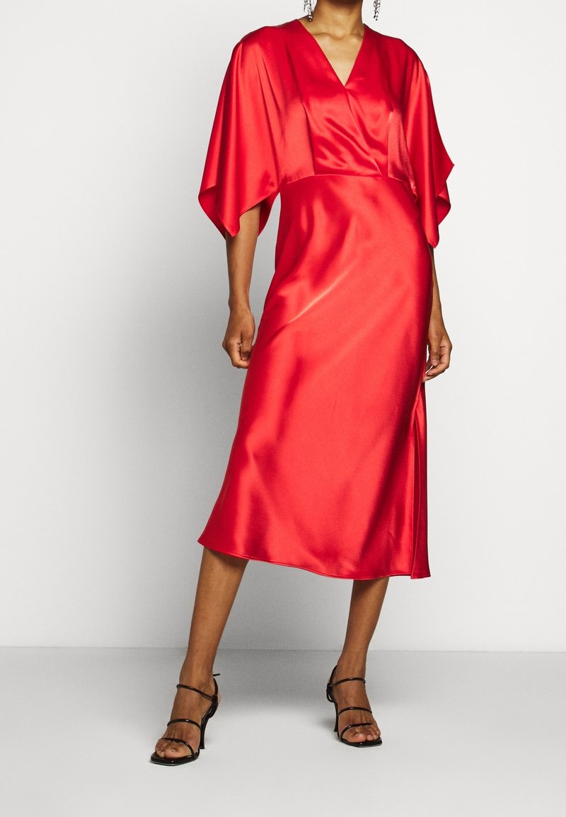 Robe en satin rouge avec un décolleté en V et des manches larges, longueur genou, texture lisse, fente latérale, portée avec des escarpins noirs à brides.