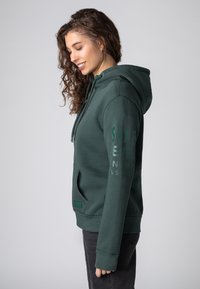 Groene hoodie van zachte stof, met een voorzak, een capuchon met trekkoord en geprinte tekst langs de mouw. Eenvoudig, casual ontwerp.