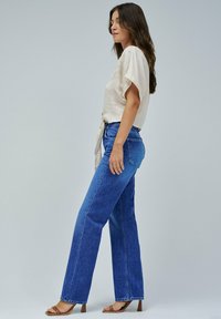 Top beige fluido con maniche corte e una cintura annodata in vita, abbinato a jeans a gamba dritta blu a vita alta e sandali con tacco marroni.