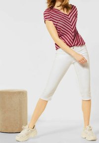 Top de manga corta a rayas en chevron rojo y blanco con escote en V, combinado con pantalones blancos de corte cropped y zapatillas beige, sobre un fondo plano.