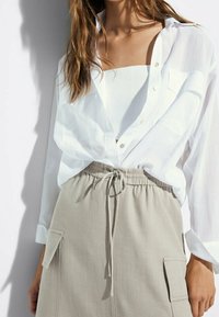 Hvid skjorte med knaplukning, let materiale, semi-gennemsigtig, lagdelt over en hvid tanktop. Beige nederdel med snøre og to sidelommer.