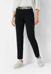 Pantalon noir slim à la texture lisse, avec une ceinture argentée, des poches latérales et un détail subtil de fermeture éclair. Porté avec des baskets blanches.