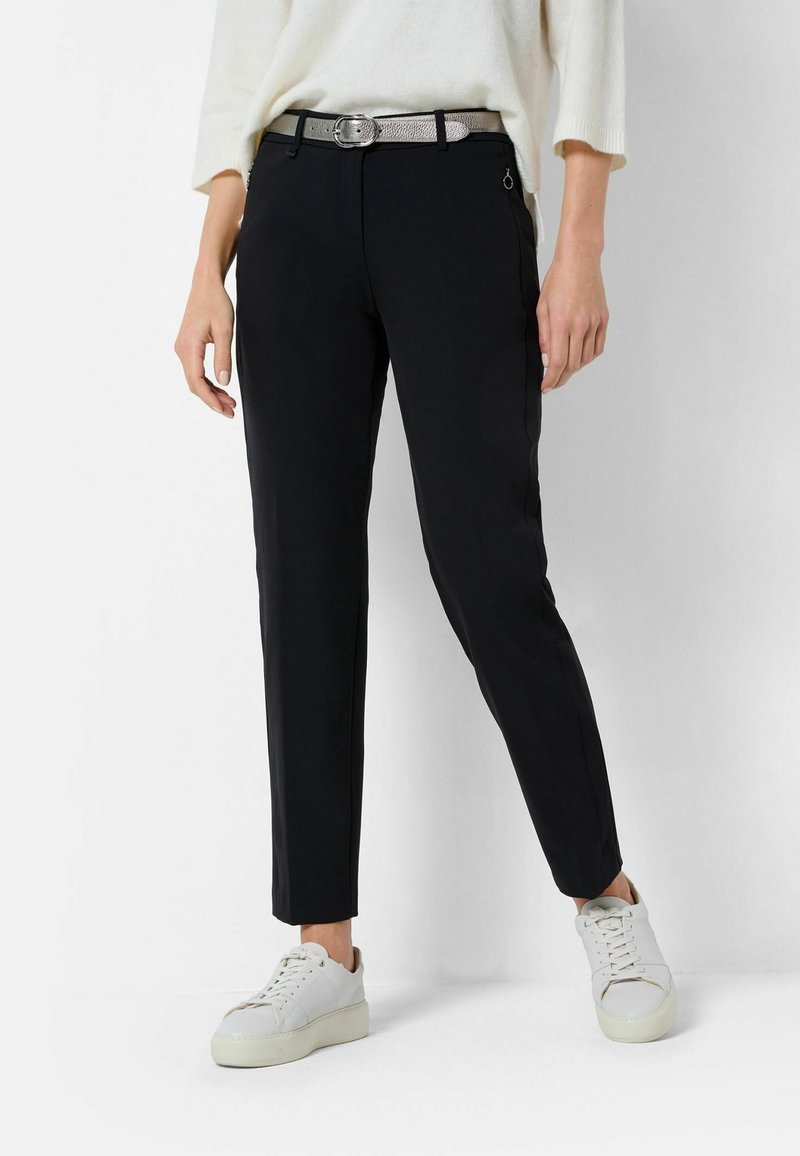 Pantalon noir slim à la texture lisse, avec une ceinture argentée, des poches latérales et un détail subtil de fermeture éclair. Porté avec des baskets blanches.
