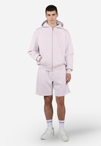 Sudadera de cremallera rosa claro con bolsillos, combinada con pantalones cortos a juego. Ambos hechos de una tela suave, con un corte relajado y puños acanalados.
