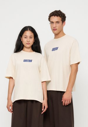 Deux mannequins se tiennent côte à côte, portant des t-shirts couleur crème avec un texte bleu "URTRN" et des pantalons amples marron foncé sur un fond uni.
