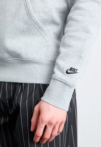 Gray cotton sweatshirt with a hood, featuring a black embroidered Nike logo on the sleeve; paired with black striped pants.