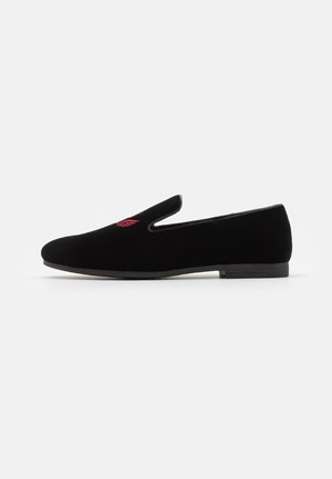 Chaussure slip-on en suede noir avec une forme incurvée, ouvertures latérales élastiques et détail de broderie rouge sur le dessus, dotée d'une semelle noire basse.