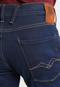 Mörkblå denimjeans med hög midja, klassiska bakfickor, en tagg i tanfärgad läder och kontrasterande gula sömnadsdetaljer.