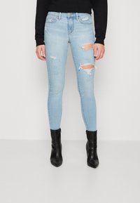 Jeans ajustados de mezclilla azul claro con detalles desgastados, presentando desgarros en ambas rodillas. Combinados con botas negras hasta el tobillo.