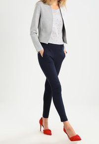 Woman wearing a light grey blazer, white patterned top, navy slim pants, and red high-heeled shoes, standing with hands in pockets.