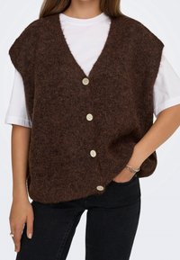 Personne portant un gilet sans manches marron foncé duveteux et boutonné sur une chemise blanche à manches courtes et un pantalon noir, main dans la poche.