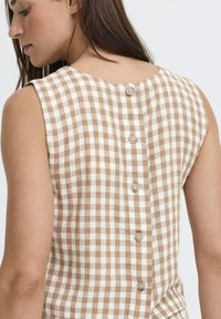 Femme portant un haut sans manches beige et blanc à carreaux vichy avec des boutons disposés verticalement dans le dos, vue de profil sur un fond blanc.