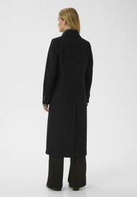 Woman with blonde hair wearing a long black coat, dark pants, and black shoes, standing facing away against a plain background.