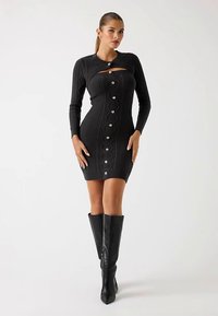 Robe noire en maille avec manches longues, dotée d'un détail découpé et de fermetures à boutons sur le devant. Associée à des bottes noires montantes jusqu'au genou.