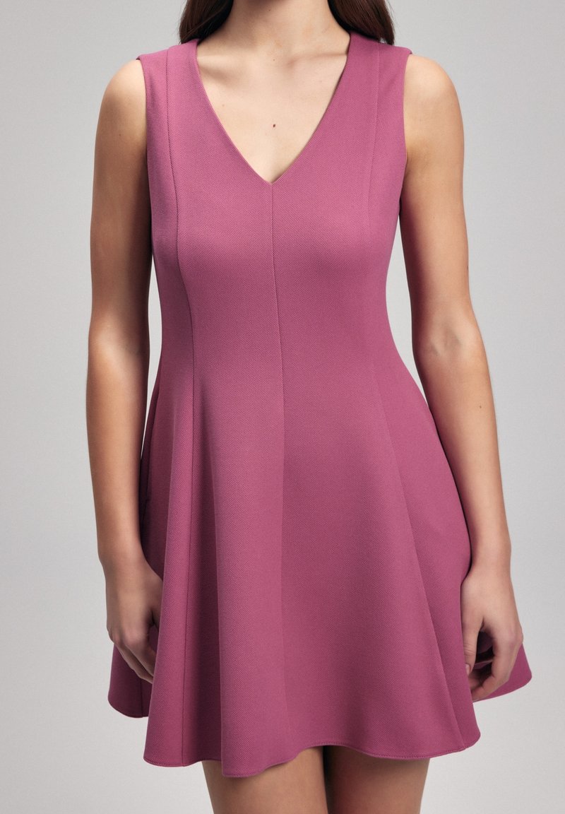 Robe sans manches à décolleté en V en tissu texturé de couleur rose moyen. Jupe évasée avec un corsage ajusté. Se termine au-dessus du genou. Détails de couture à l'avant.