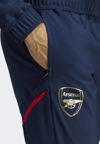 Marineblauwe sportbroek met elastische tailleband, voorzien van een gouden Arsenal-logo en een rode ritszak voor extra gemak. Glad van structuur.