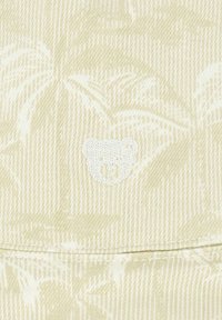 Tessuto crema con un sottile motivo di foglie di palma, con un piccolo volto di orsetto ricamato in bianco centrato sotto. Texture liscia.