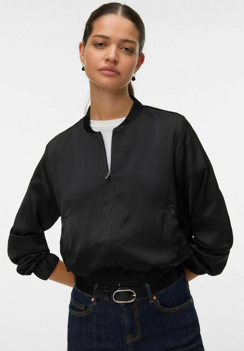 Giacca bomber nera con una texture liscia, simile a satin, dotata di chiusura con zip, colletto alto e vita elasticizzata. Indossata con una camicia bianca.