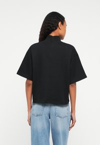 Frau mit langen, lockigen Haaren, die ein schwarzes kurzärmliges Shirt mit Stehkragen und hellblaue hochtaillierte Jeans trägt, steht mit dem Rücken zur Kamera.