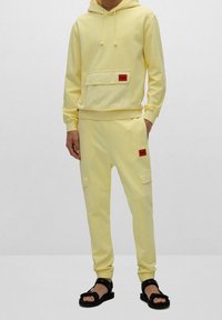 Felpa gialla in cotone con tasca frontale, abbinata a pantaloni jogger coordinati con tasche laterali e accenti di logo rossi. Sandali neri completano il look.