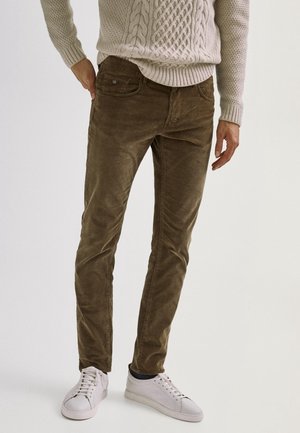 Mężczyzna w brązowych spodniach ze sztruksu, beżowym swetrze w warkocze i białych trampkach, stojący na tle jasnego, jednolitego tła.