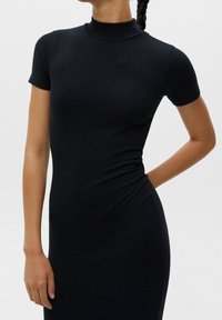 Femme portant une robe noire moulante à manches courtes et col roulé, avec une tresse sur l'épaule, debout devant un fond uni.