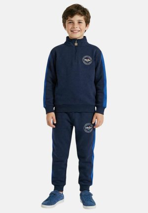 Garçon debout, souriant, portant un survêtement bleu marine avec des rayures bleu clair et des baskets assorties, sur un fond uni.