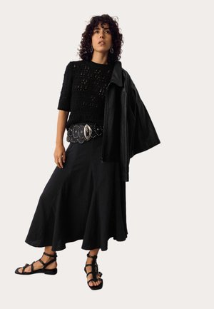 Vrouw met krullend haar, gekleed in een zwart gehaakt topje, een brede riem, een lange zwarte rok, gladiatorsandalen en een leren jas over één schouder gedrapeerd.