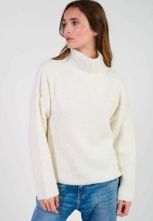 Pull à col roulé de couleur crème en matériau texturé et duveteux, avec des épaules tombantes et des manches longues, associé à un jean bleu clair.