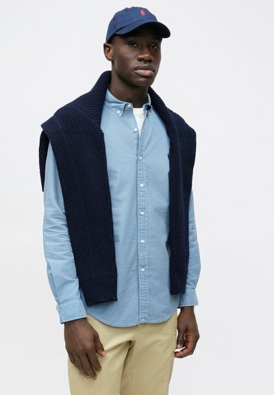 Colete de malha azul-marinho usado por cima de uma camisa azul-claro com botões. Calças bege claro. Boné azul com emblema vermelho. Tom de pele escuro. Fundo simples.