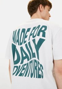 Witte katoenen t-shirt met in het oog springende groene grafische tekst op de rug: "MADE FOR DAILY ADVENTURES." Korte mouwen, relaxte pasvorm.
