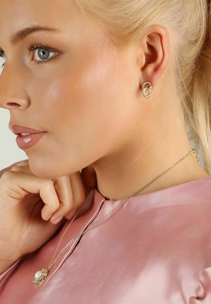 Guldörhängen med sammanflätade cirkulära accenter; modellen bär en ljusrosa satintopp och ett delikat guldhalsband med en pärldetalj.