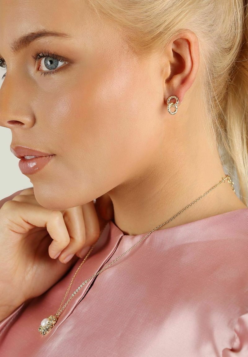 Guldörhängen med sammanflätade cirkulära accenter; modellen bär en ljusrosa satintopp och ett delikat guldhalsband med en pärldetalj.