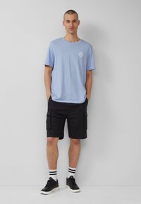 Helles blaues Baumwoll-T-Shirt mit Druck auf der Brust, kombiniert mit schwarzen Cargo-Shorts, schwarzen Turnschuhen und schwarz-weiß gestreiften Socken.