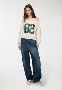 Felpa oversize crema con grafica verde "82", abbinata a jeans di denim blu a gamba larga e sneakers beige. Texture liscia e vestibilità comoda.