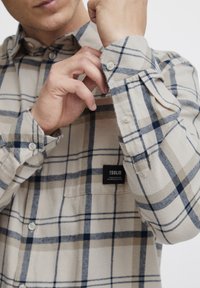 Camicia a quadri beige e blu navy realizzata in tessuto morbido, con colletto botton-down, polsini con bottoni e un piccolo etichetta del marchio sul petto.