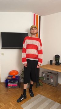 Czerwono-biały sweter z paskami, wykonany na drutach, w luźnym kroju, zestawiony z czarnymi szortami do kolan i czarnymi sportowymi butami. W tle znajduje się telewizor i dekoracje.