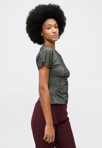 Mulher com cabelo afro, vestindo uma blusa cinza justa de mangas curtas e calças bordeaux de cintura alta, em pé contra um fundo claro liso.